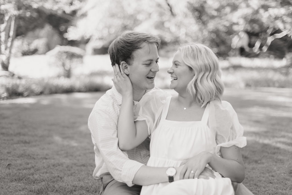 Couple sits in a shaded, grassy area. Female is sitting in her fiance's lap with her hand on his cheek as they laugh and smile at each other. This is a fun pose example for a Birmingham Engagement Session